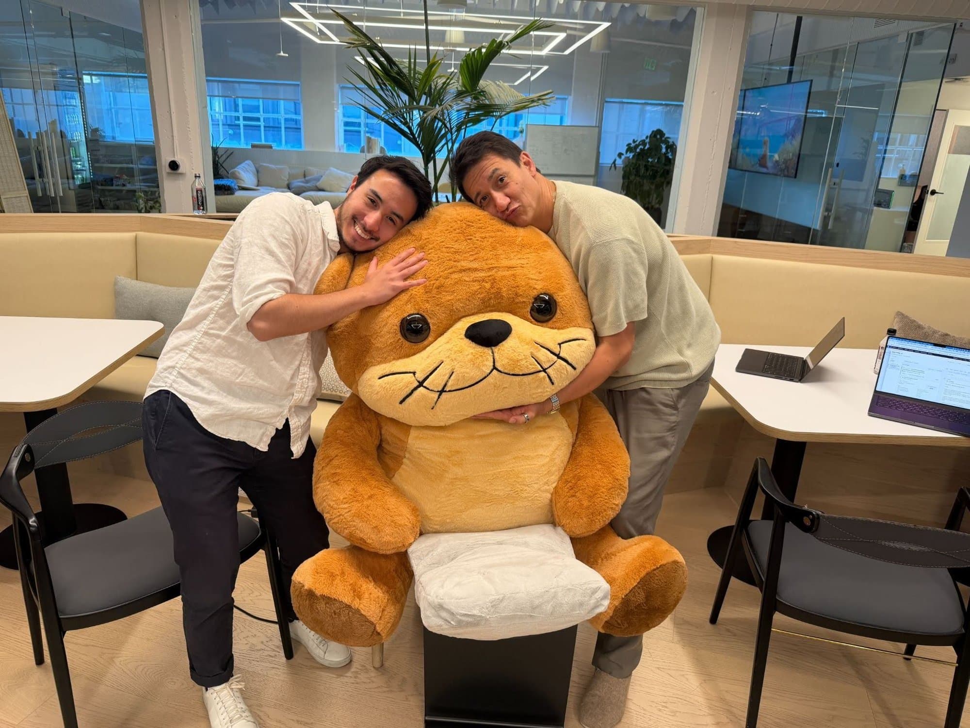 Alex Dengler and a teammate hugging a giant stuffed otter in the office